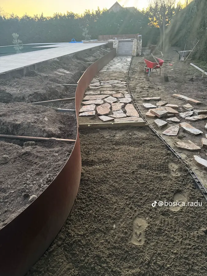 Bordură curbată din oțel corten lângă o alee din piatră naturală, fotografiată la apus.
