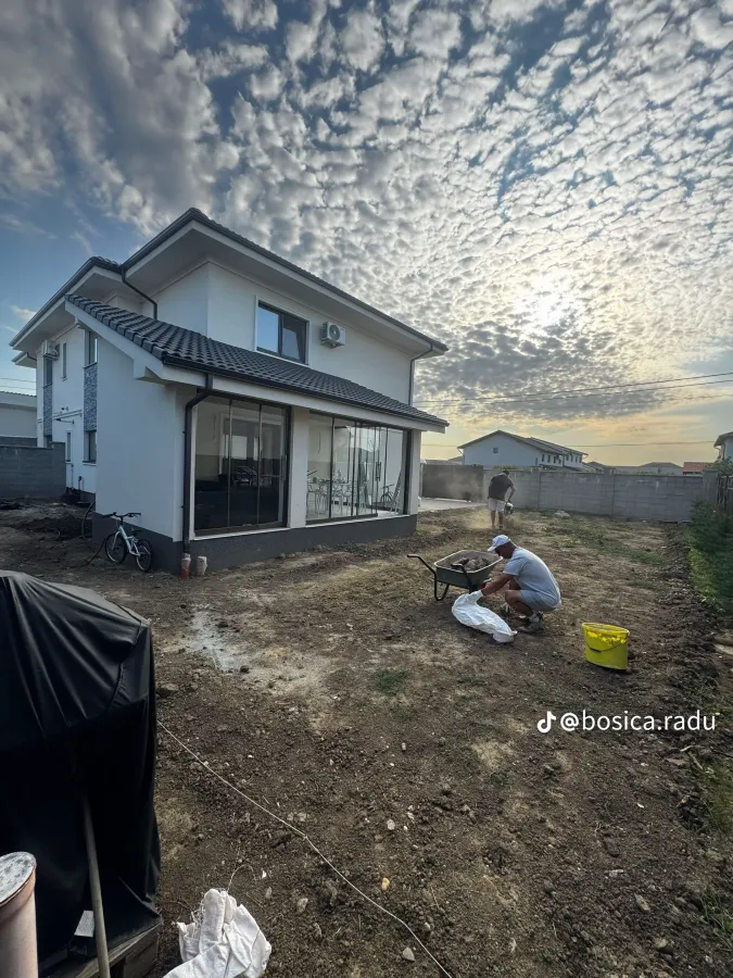 Doi muncitori pregătesc terenul unei curți noi, la asfințit, înainte de montarea gazonului.